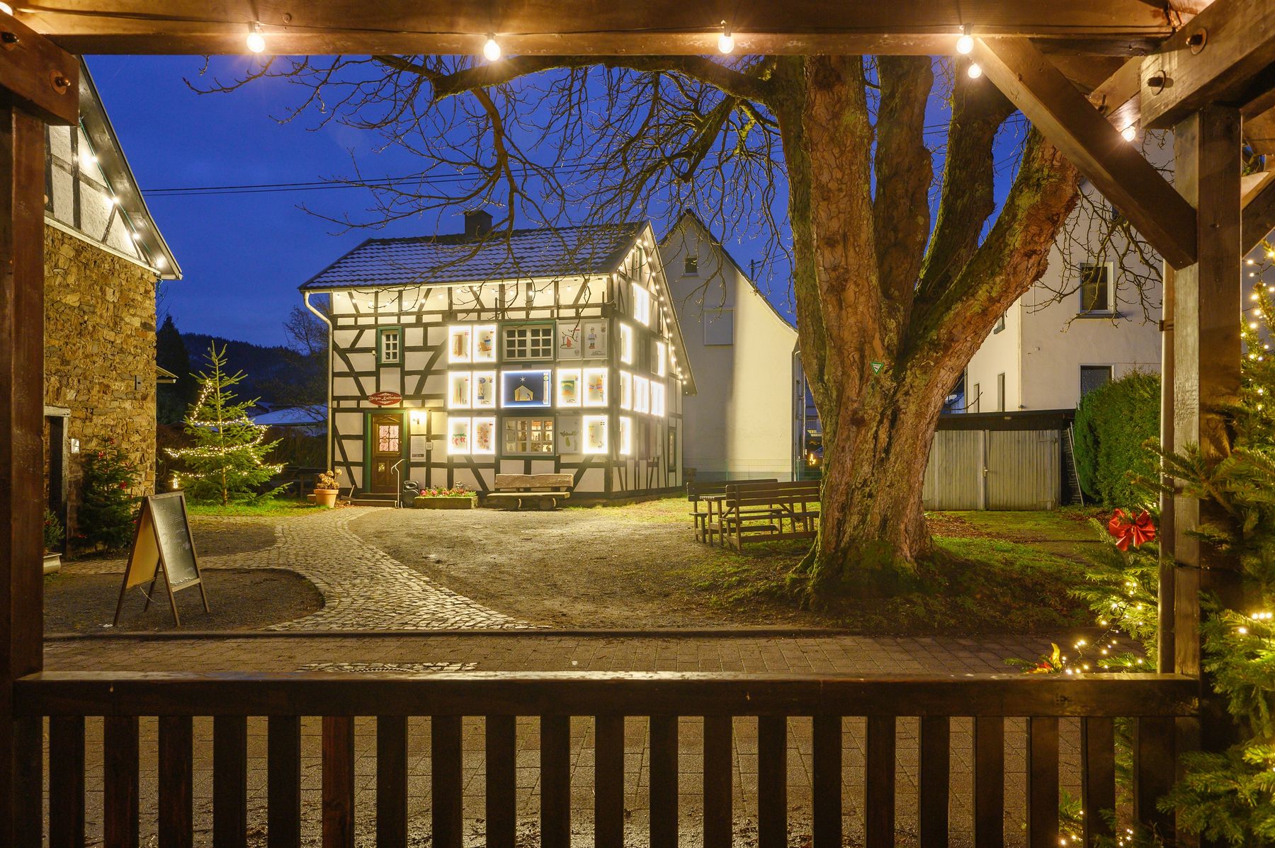 A half-timbered house with glowing windows is framed by trees, benches, and festive string lights on a quiet, cobblestone courtyard at dusk.